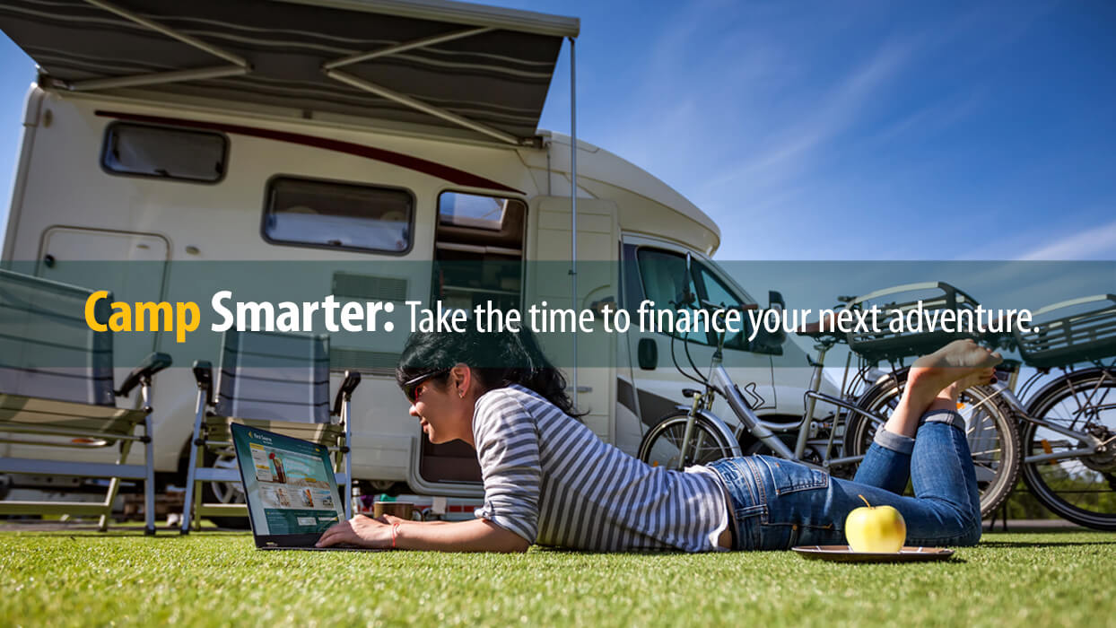 Young woman lying on ground using laptop at campsite with camper financed through First Source